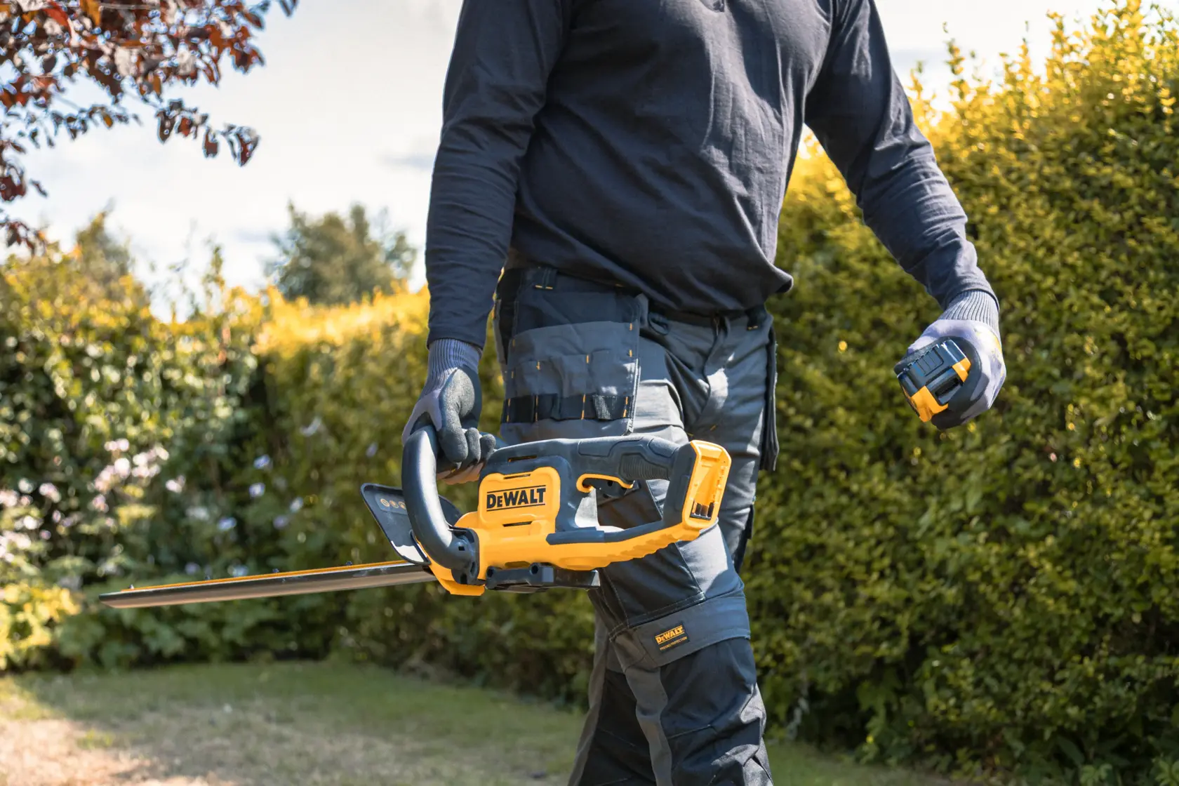 Person holding a DEWALT cordless hedge trimmer outdoors, wearing gloves and work pants in a garden.
