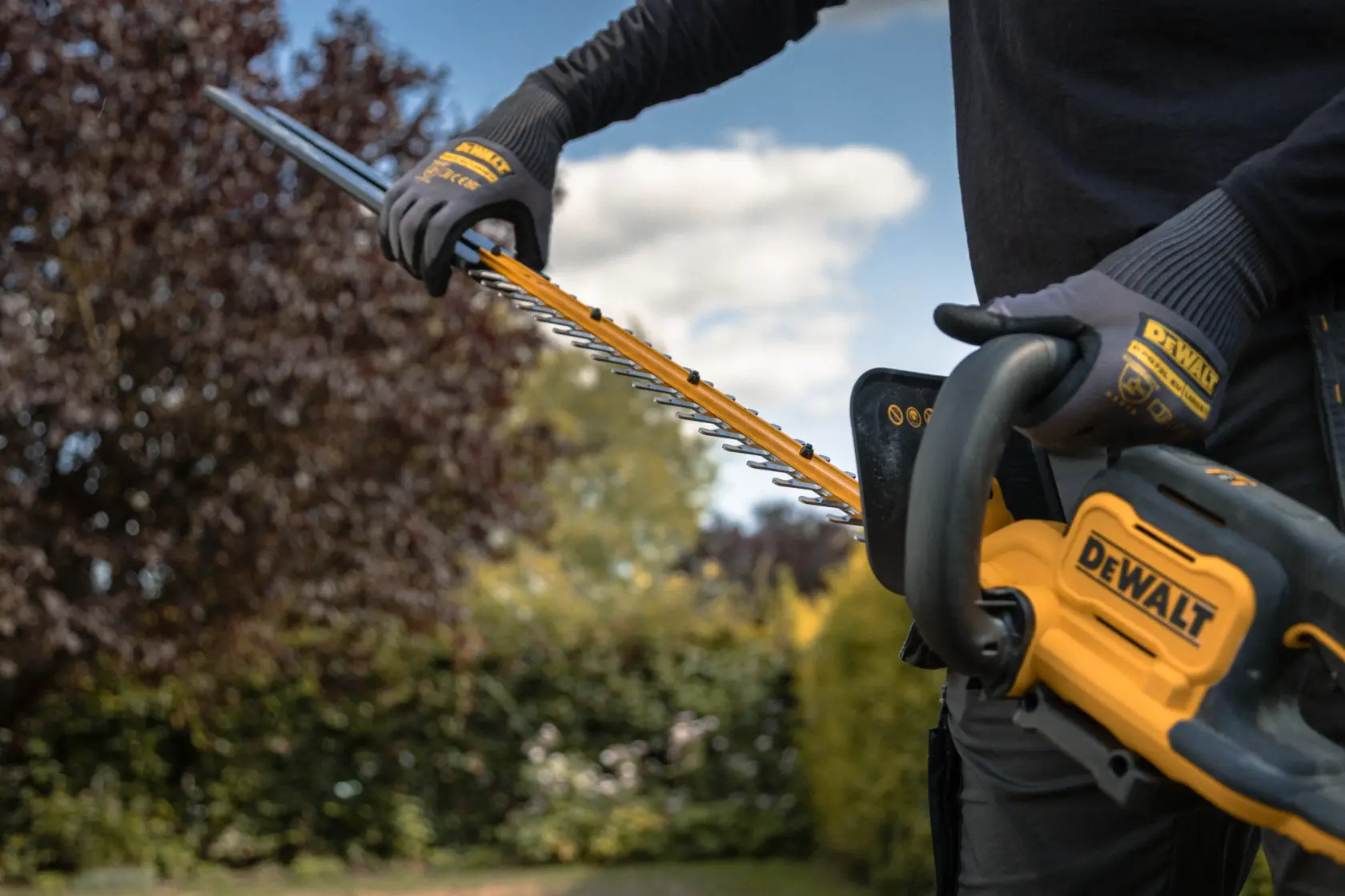 Person using a DEWALT cordless hedge trimmer outdoors with gloves on, surrounded by garden plants.