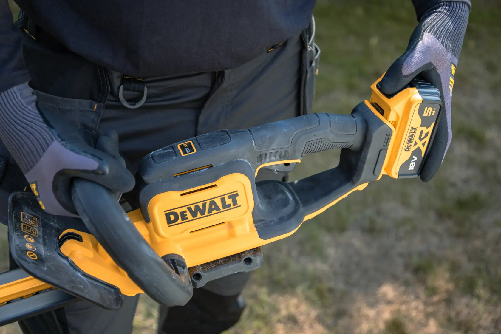 Close-up of gloves holding a DEWALT cordless hedge trimmer outdoors with visible battery pack.