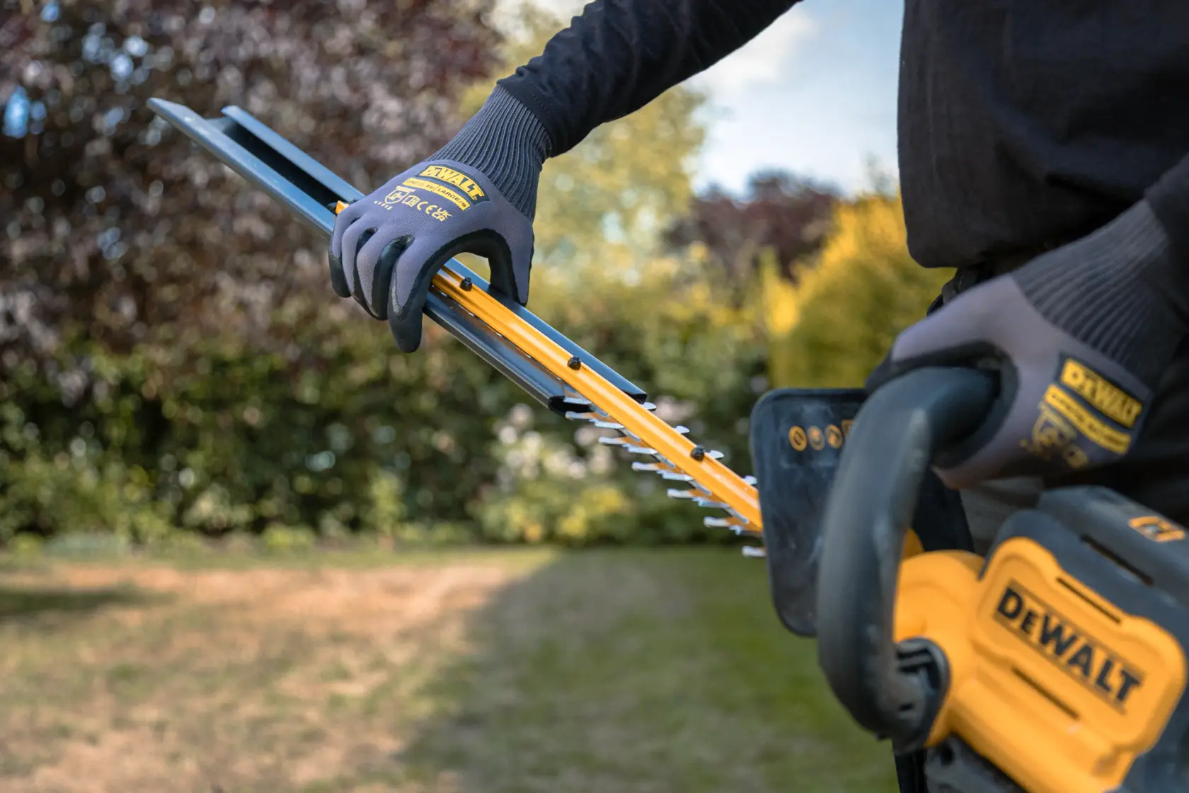 Person using a DEWALT cordless hedge trimmer in a garden, wearing protective gloves.