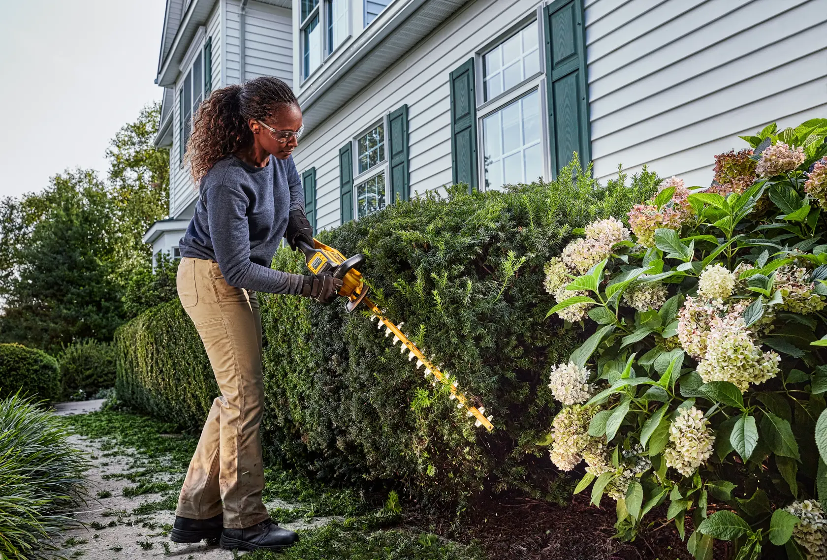 Person trimmt Büsche mit einem DEWALT Akku-Heckenschneider im Garten vor einem Haus.