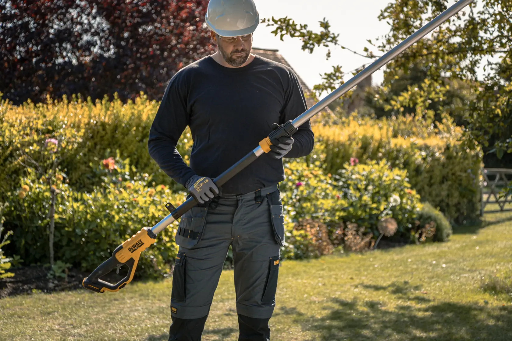 Person holding a DEWALT cordless pole saw outdoors with garden plants in the background.