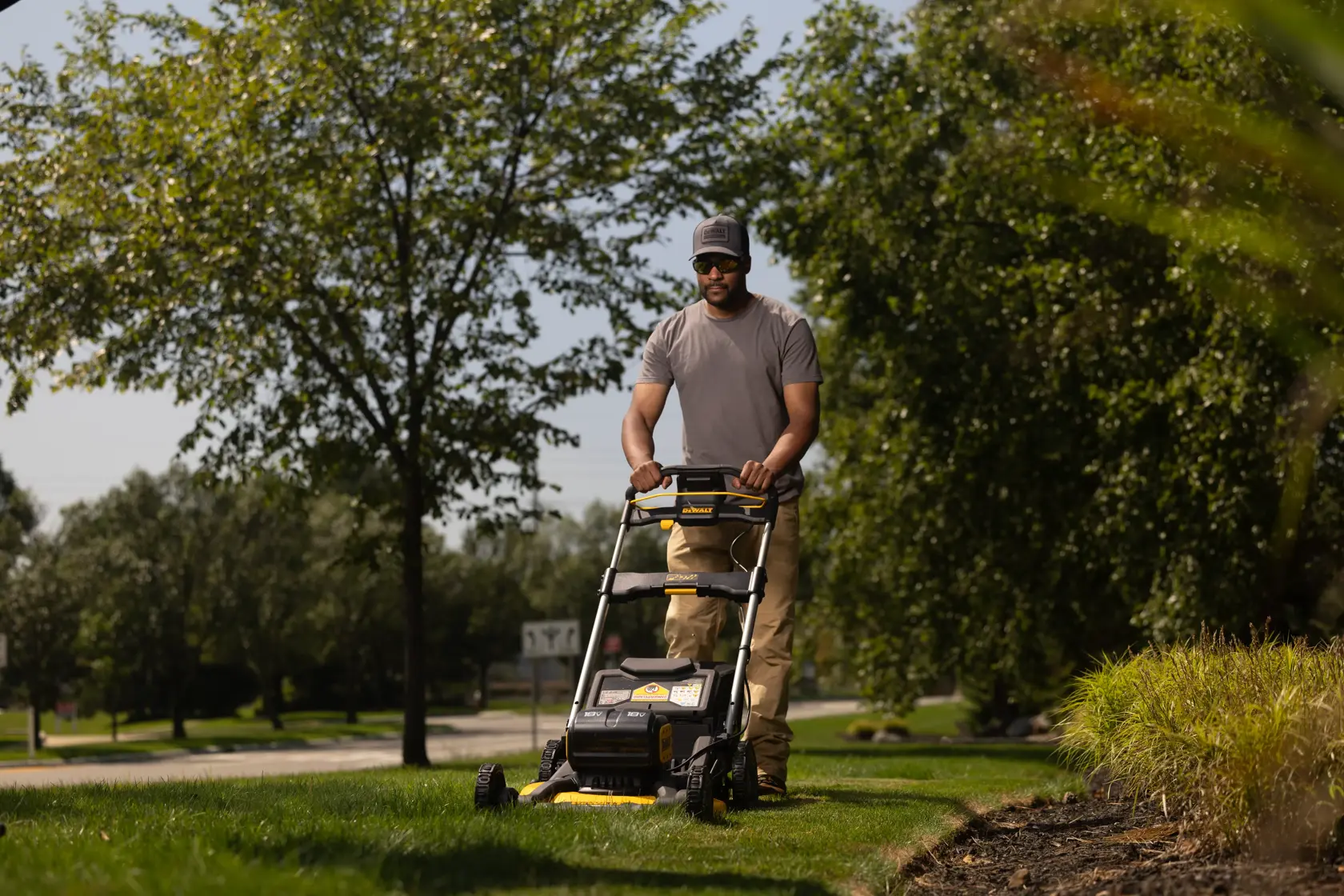 A person mowing a lawn with a DEWALT cordless lawn mower model DCMWP134N, surrounded by green trees and plants on a sunny day.