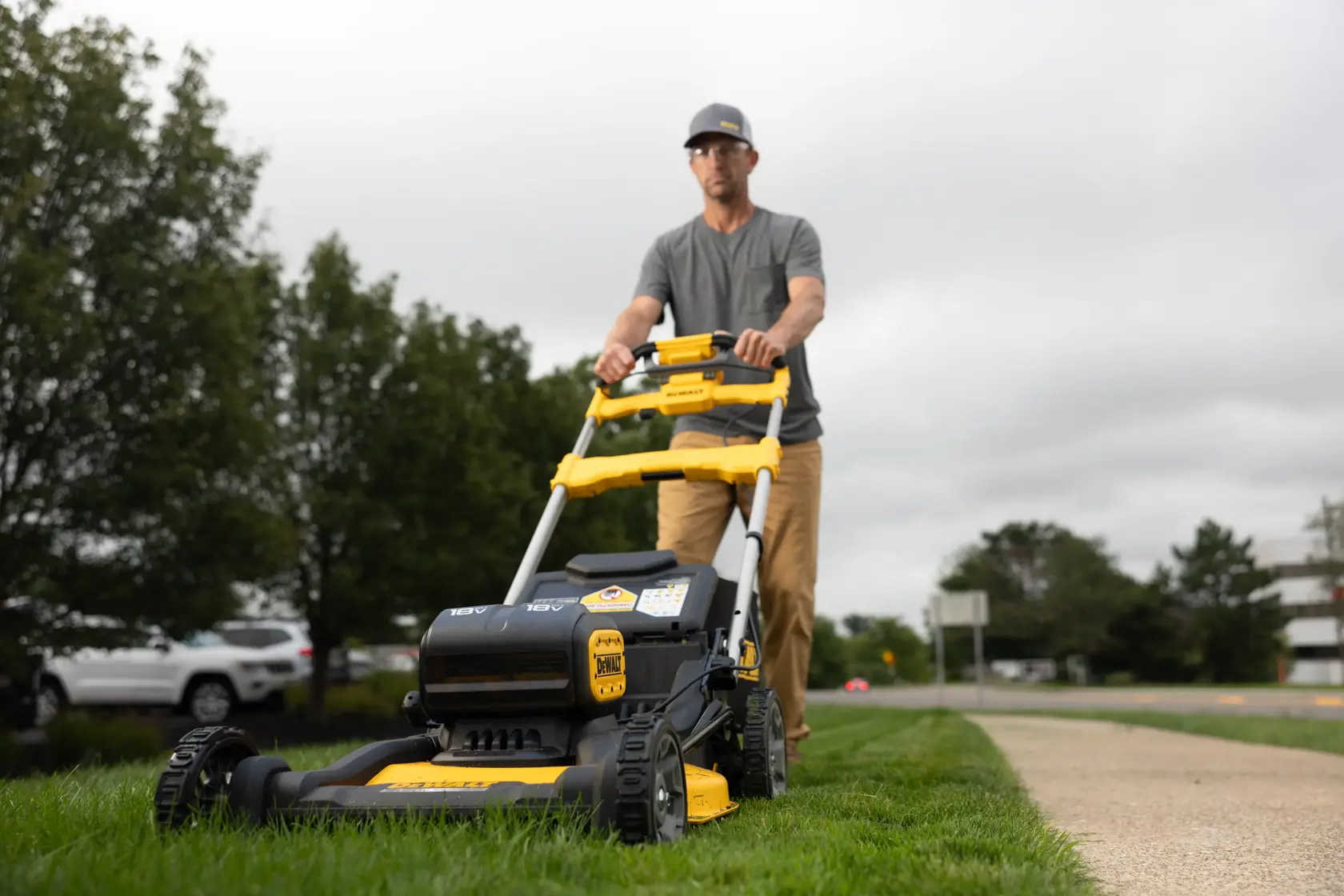 A person is pushing a DEWALT lawn mower on grass next to a sidewalk, with trees and parked cars in the background. The person's face is blurred.