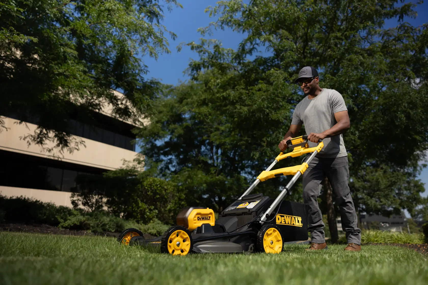 Person mäht mit einem DEWALT Rasenmäher draußen auf Gras, Gebäude und Bäume im Hintergrund.