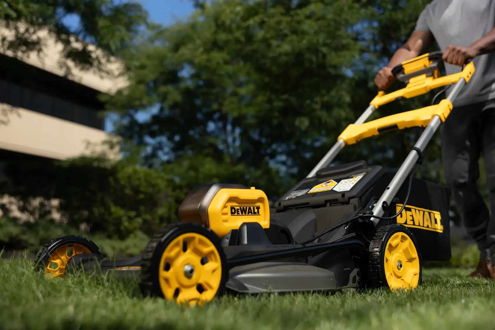 Ein DEWALT Rasenmäher wird draußen auf Gras geschoben, mit Bäumen und Gebäude im Hintergrund.