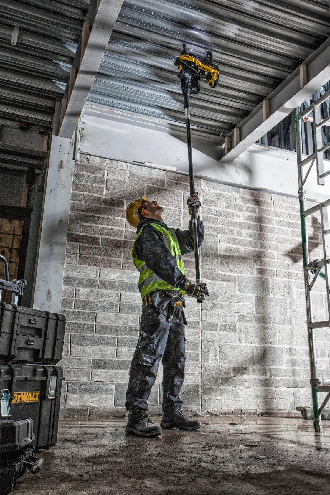 Worker uses DEWALT DCN890P2 tool overhead at construction site. Safety gear and tool cases visible.