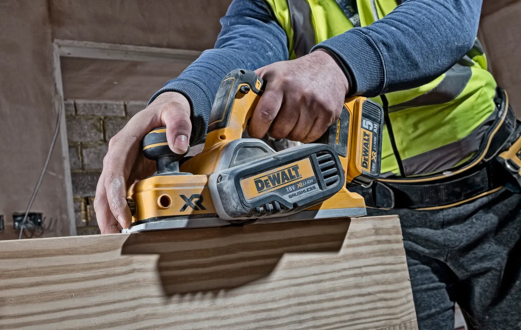 Worker uses a DEWALT cordless planer on a wooden plank in a construction setting.