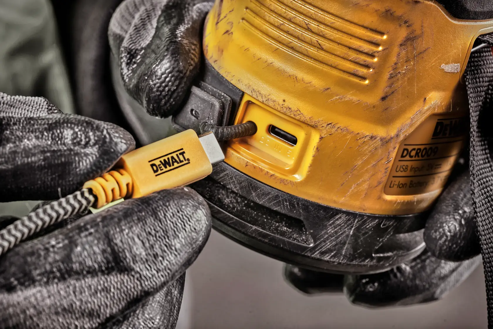 Nærbilde av en person med svarte hansker som kobler en gul DEWALT USB-C ladekabel til en DEWALT DCR009-enhet. Enheten har synlige riper og en produkttagg.