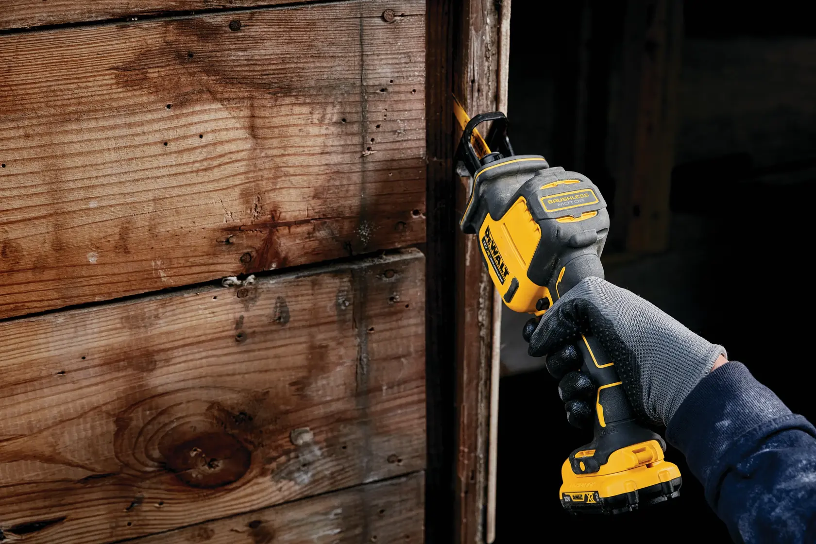 Person uses a DEWALT reciprocating saw to cut wood near a wall. Gloved hand holds the yellow and black tool.