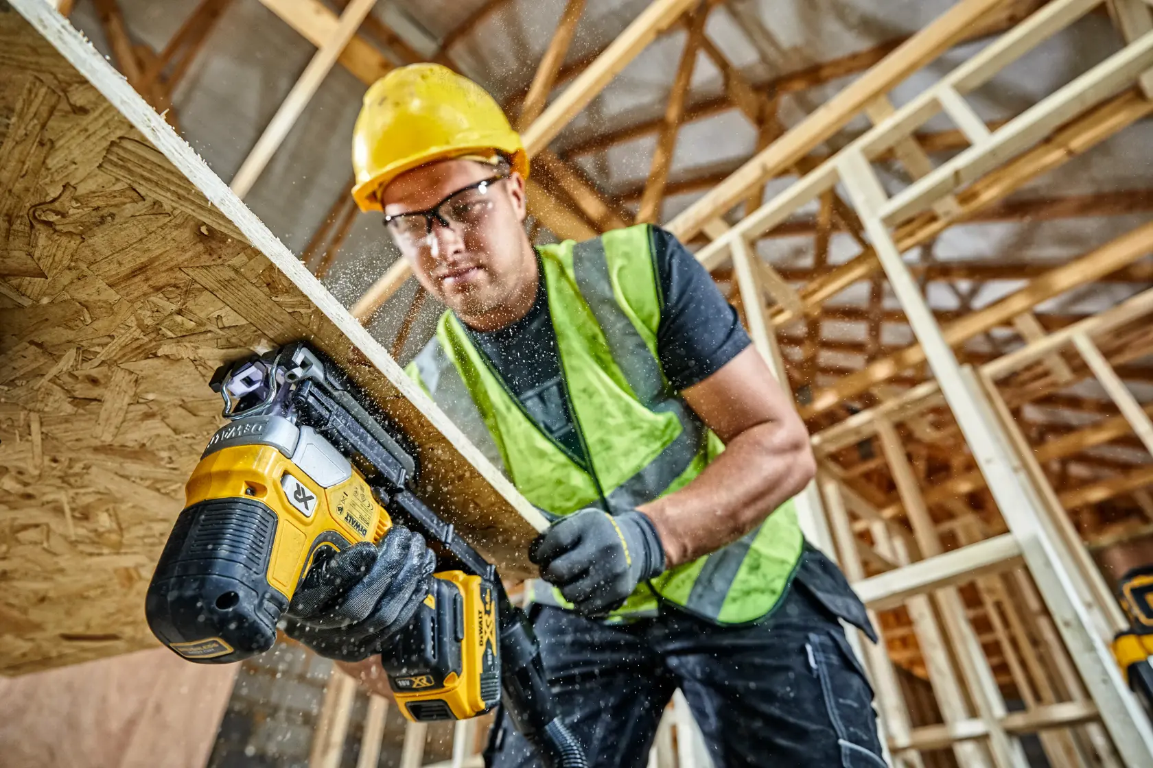 Worker using a DEWALT cordless jigsaw to cut wood at a construction site wearing safety gear.