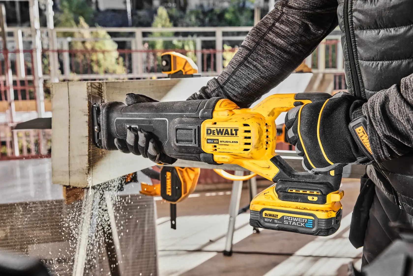 A person using a DEWALT cordless reciprocating saw to cut through a wooden beam, with sawdust flying. The saw features a DEWALT POWERSTACK battery and the scene takes place at a worksite.