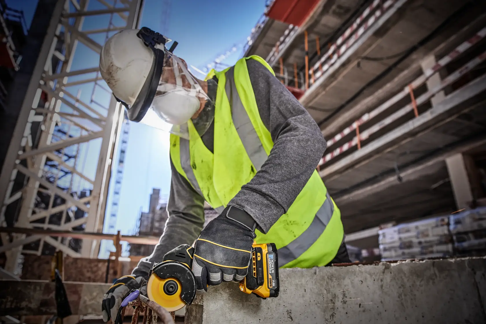 A worker wearing a safety helmet, face shield, mask, high-visibility vest, and DEWALT-branded gloves uses a DEWALT DCS438E2T cordless tool to cut through concrete at a construction site.