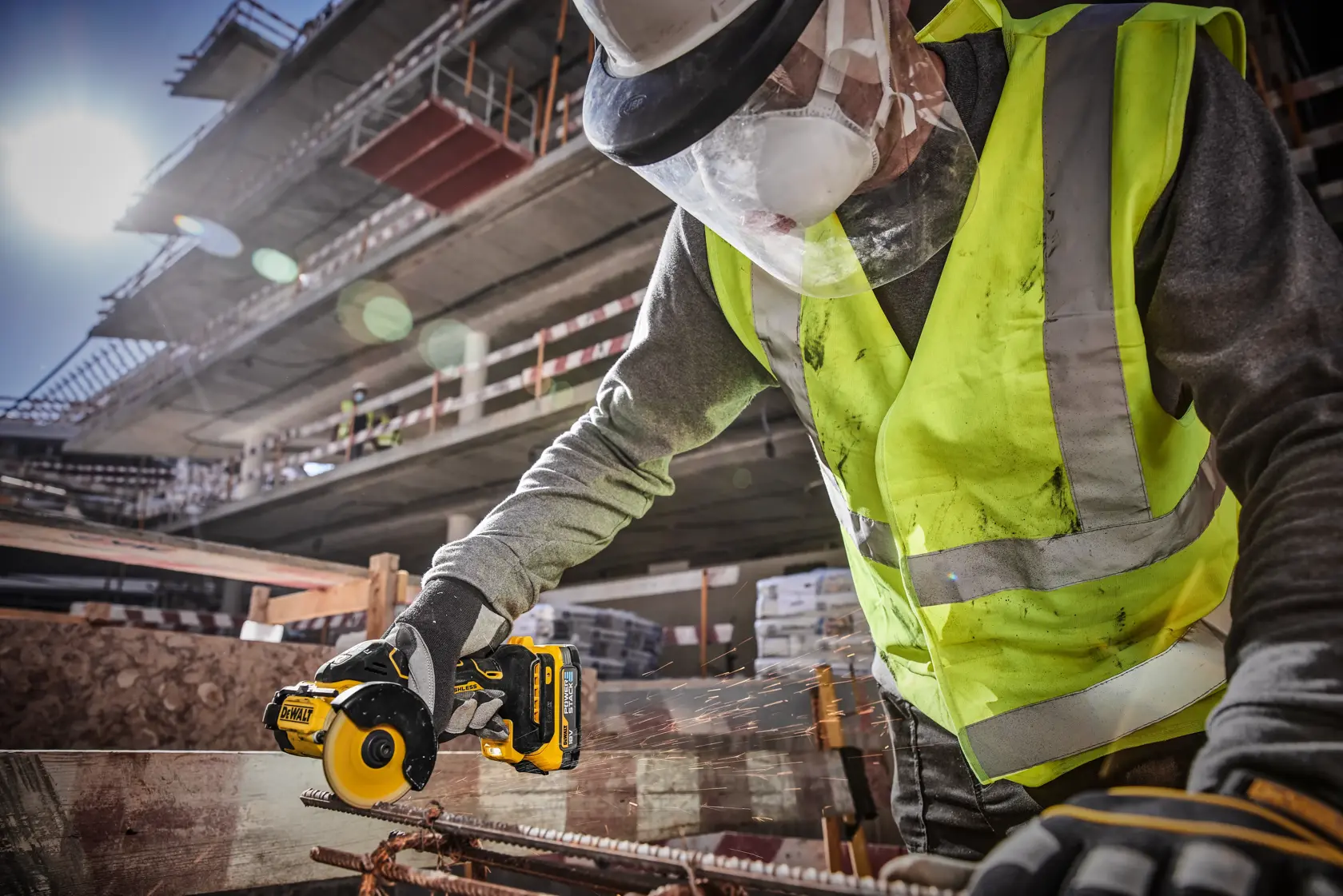 A worker wearing a hard hat, face shield, gloves, and a high-visibility vest uses a DEWALT cordless angle grinder (model DCS438E2T) on a concrete slab at a construction site.