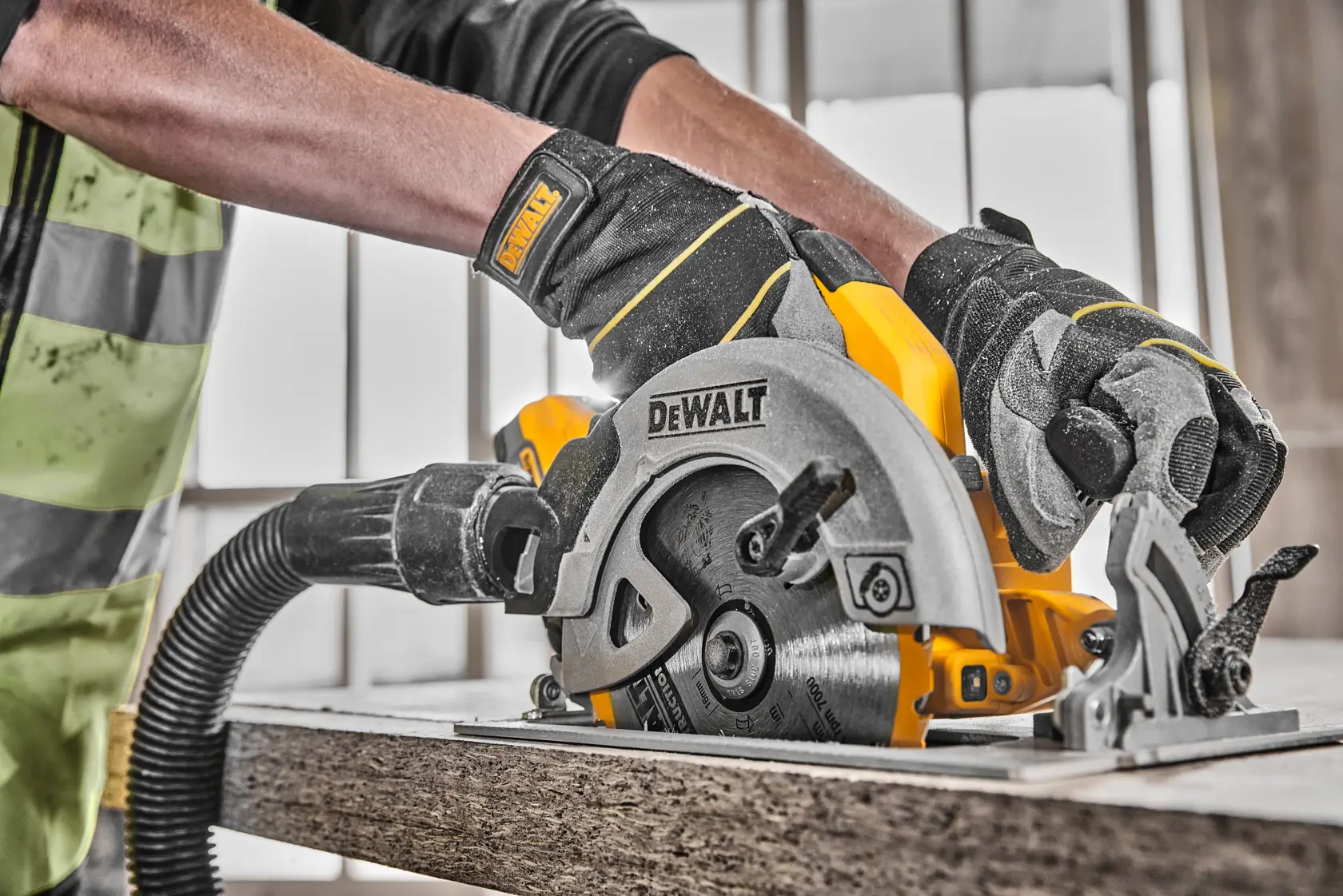 Gloved hands using a DEWALT cordless circular saw to cut wood, with a vacuum attachment.