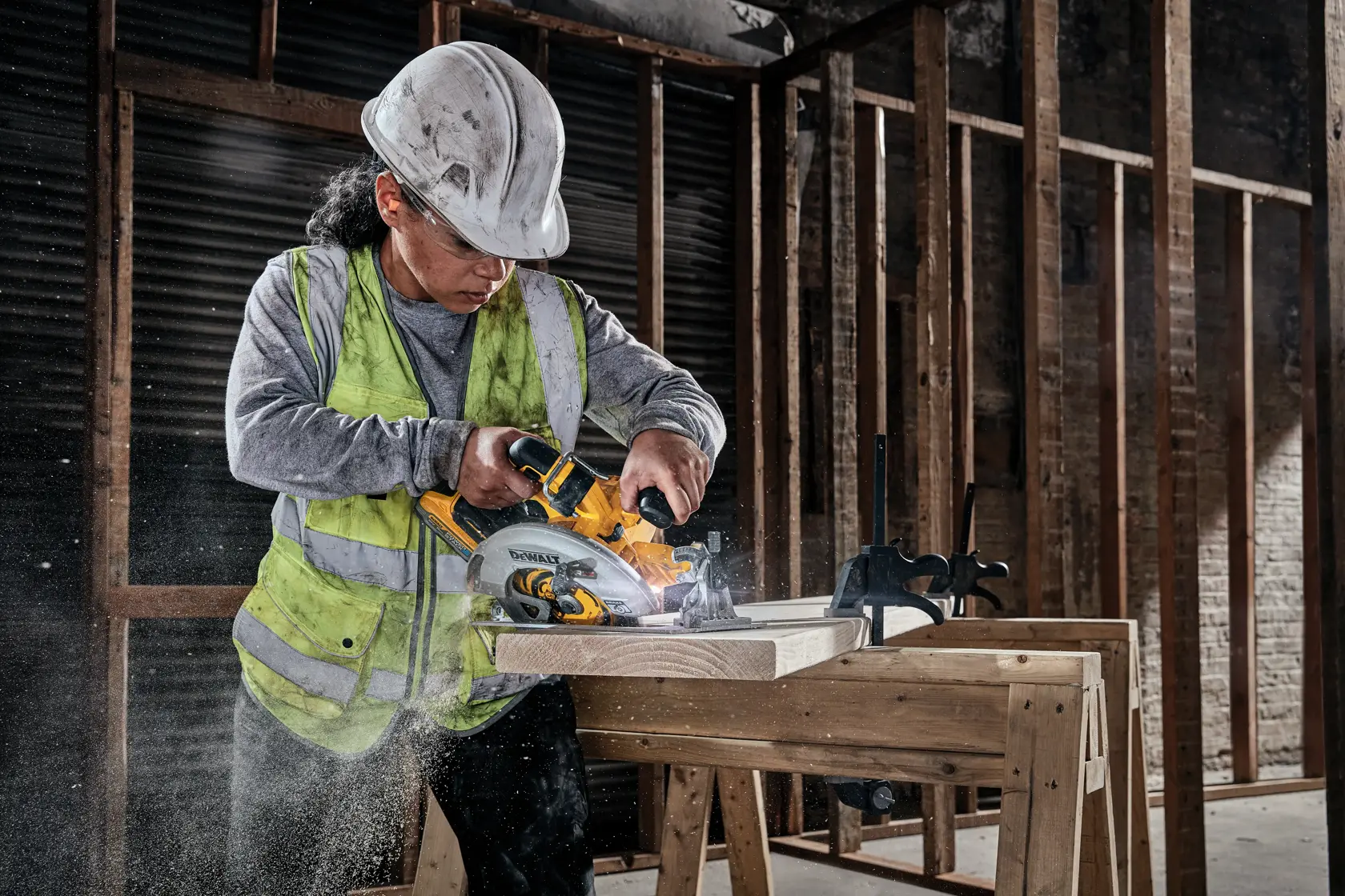 Ein Bauarbeiter mit Sicherheitshelm und Warnweste schneidet mit einer kabellosen DEWALT Kreissäge Holzlatten auf einer Werkbank in einem unfertigen Gebäude.