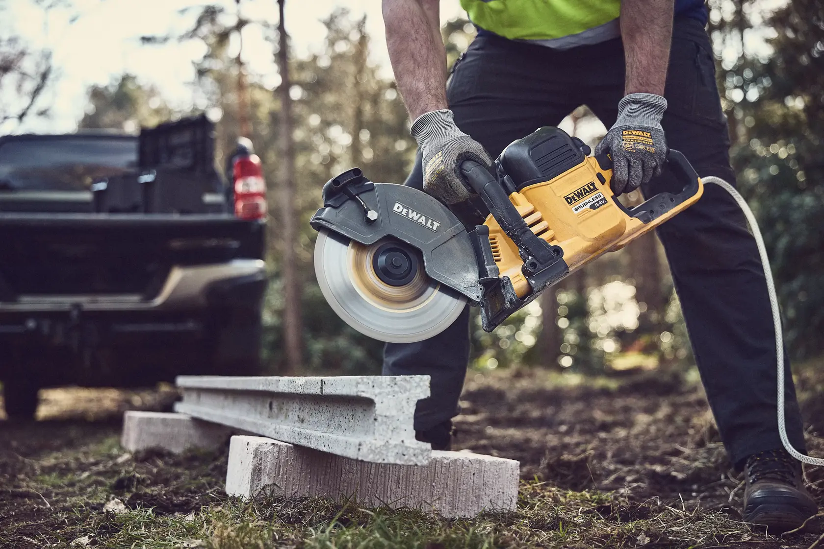 A person uses a DEWALT power saw to cut concrete outdoors. Safety gloves and a truck are visible in the background.