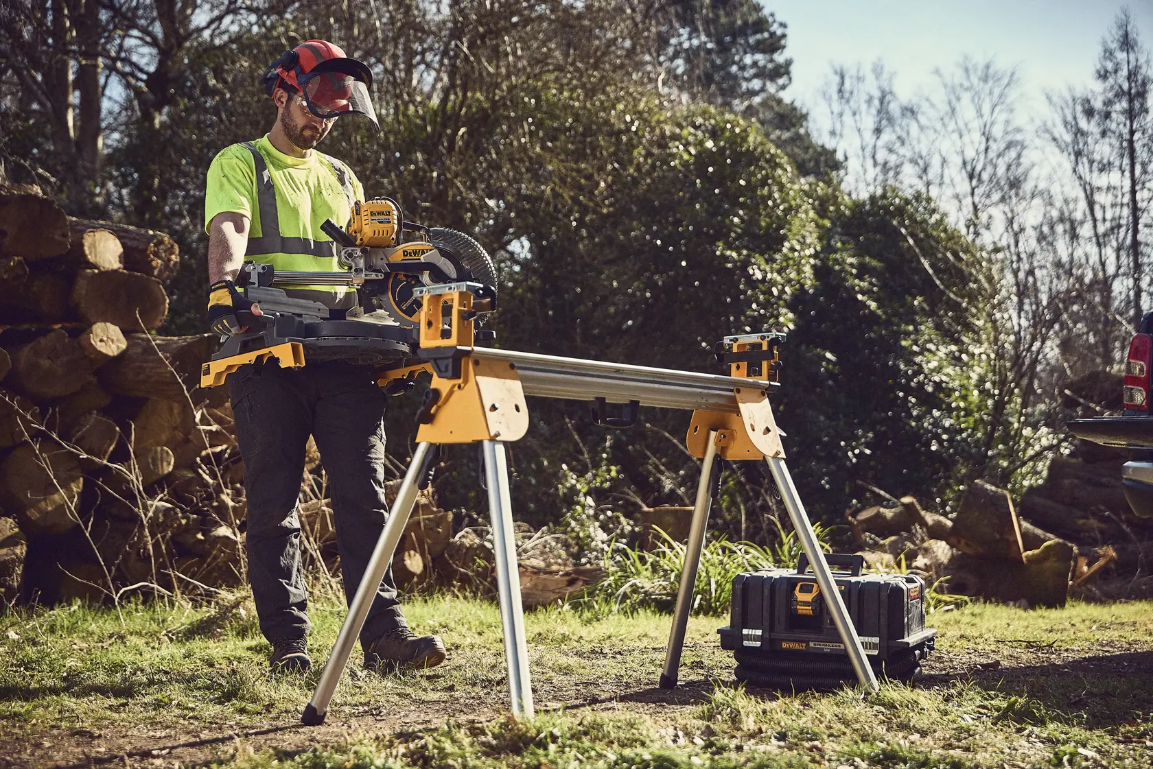 Eine DEWALT Kappsäge auf einem Ständer draußen, ein Arbeiter mit Schutzkleidung steht bei Holz und Werkzeugkiste.