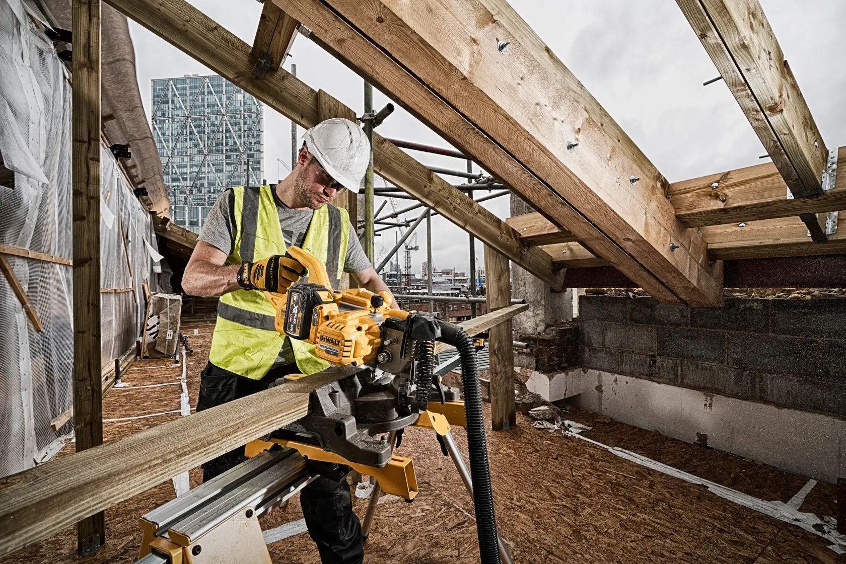 Ein Bauarbeiter mit Warnweste und Schutzhelm bedient eine kabellose DEWALT Gehrungssäge (SKU: DCS777N), um einen Holzbalken auf einer Baustelle zu schneiden. Die Umgebung zeigt teilweise errichtete Strukturen mit freiliegenden Balken und Stadtgebäude im Hintergrund.