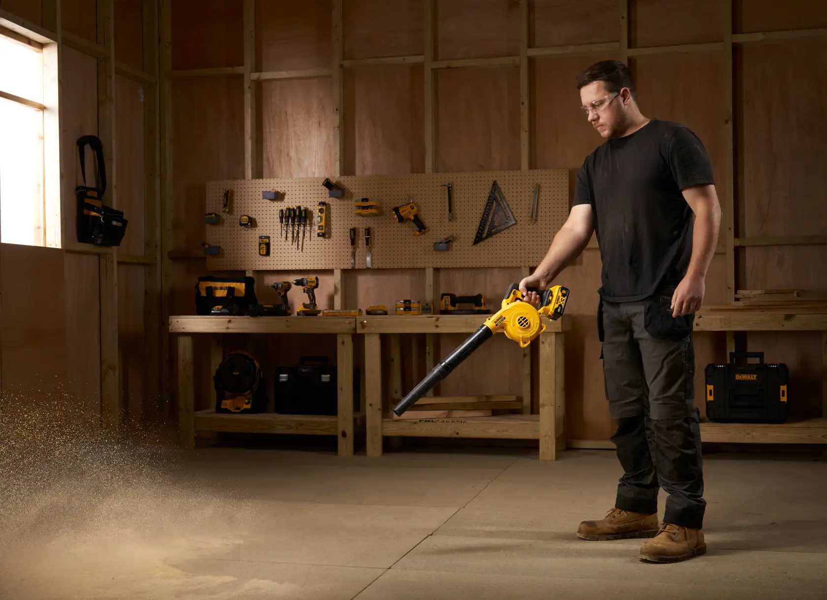 Person using a DEWALT DCV100 blower in a workshop, blowing dust from the floor. Tools visible on benches and walls.