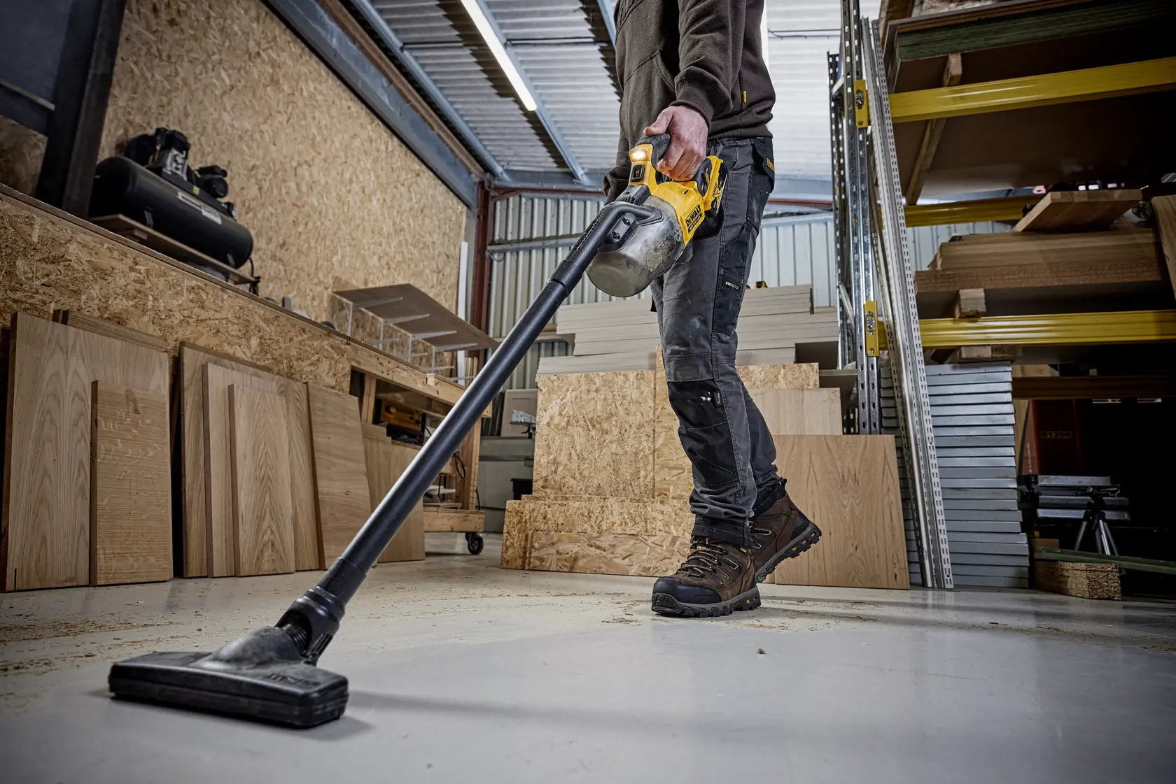 Person using a DEWALT cordless vacuum to clean a workshop floor with wooden boards and shelves visible.