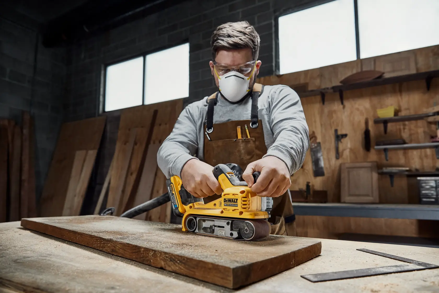 A person in a workshop uses a DEWALT DCW220N yellow belt sander on a wooden plank. The person’s face is blurred. The surroundings show wooden panels and various tools.