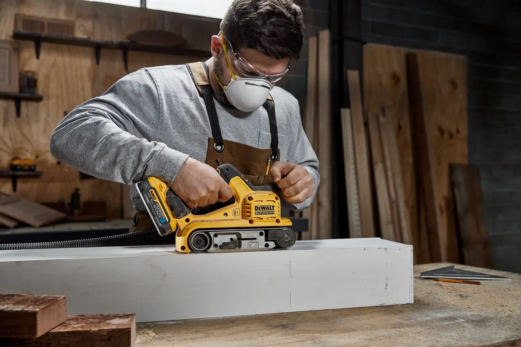 A person wearing an apron is using a yellow DEWALT DCW220N cordless belt sander to sand a large piece of wood in a workshop.