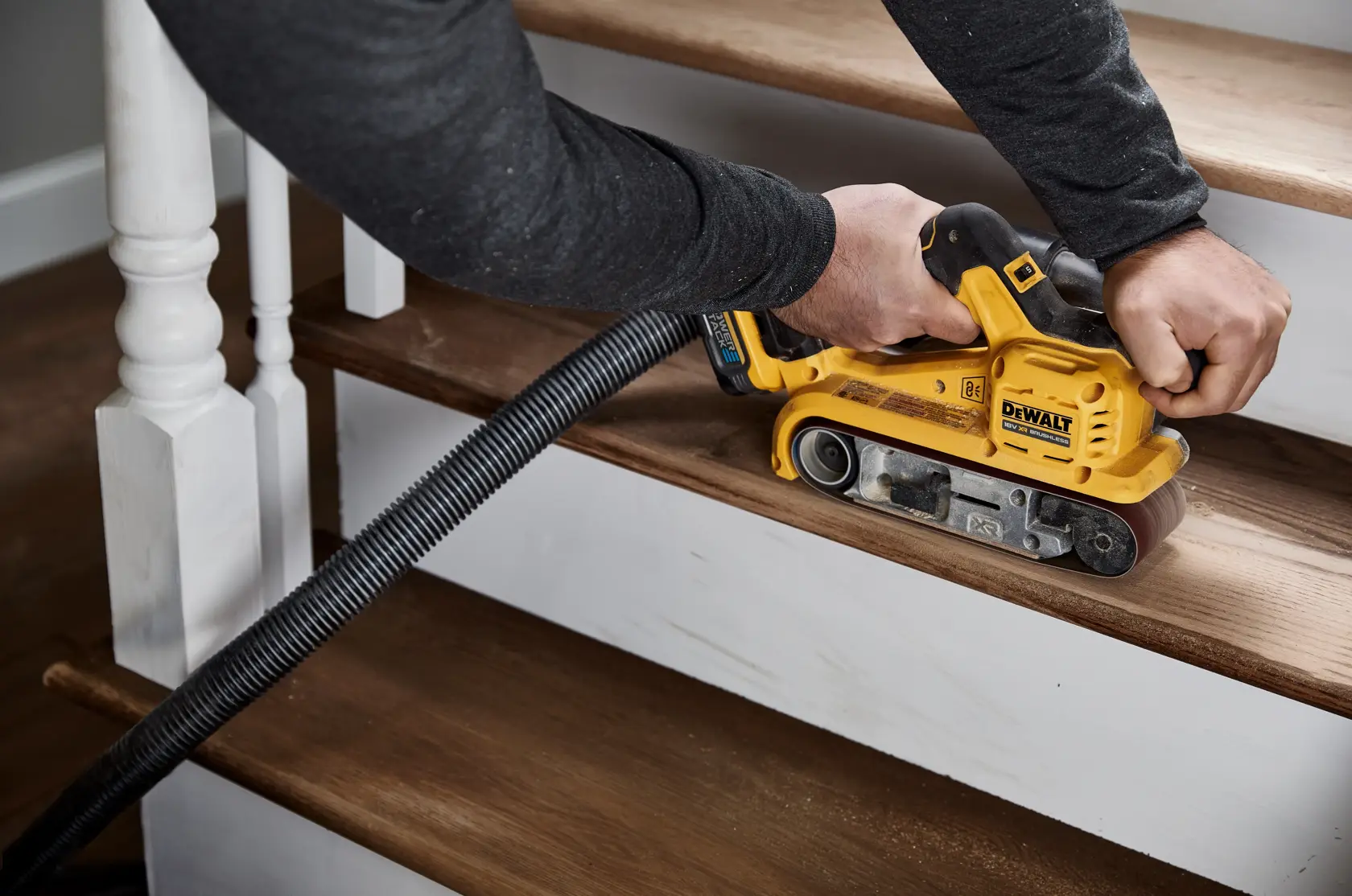 A person using a DEWALT DCW220N cordless belt sander to sand wooden stairs, with a vacuum hose attached to the tool for dust collection.