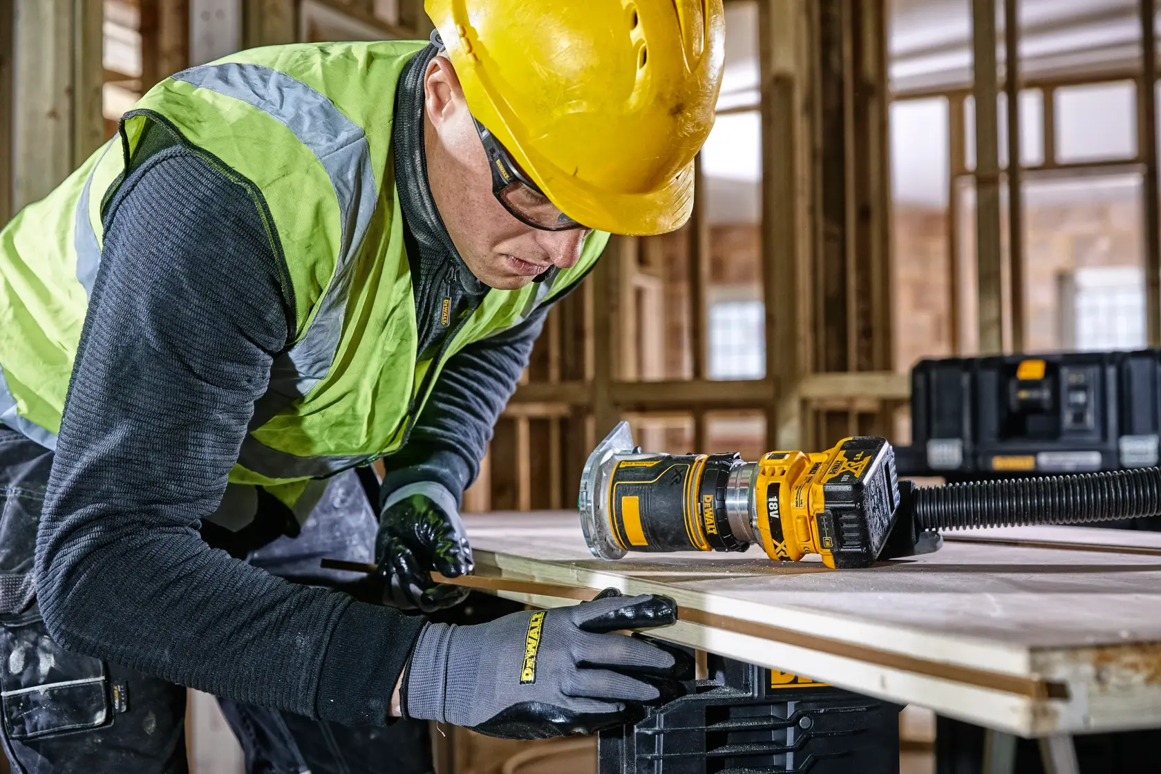 Trabajador usa la fresadora DEWALT DCW604 en madera en obra, con guantes, casco y chaleco de seguridad.