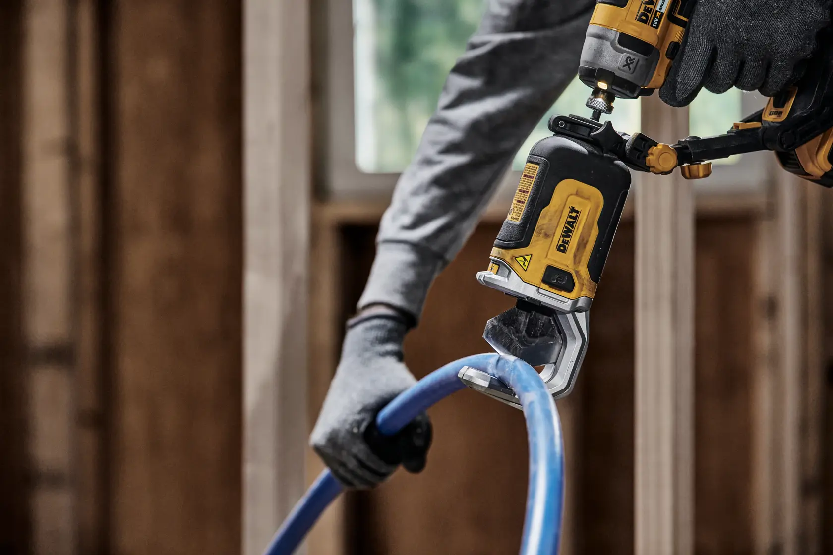 En person med hansker bruker et DEWALT DT20560 elektrisk verktøy til å kutte et blått plastrør på en byggeplass.