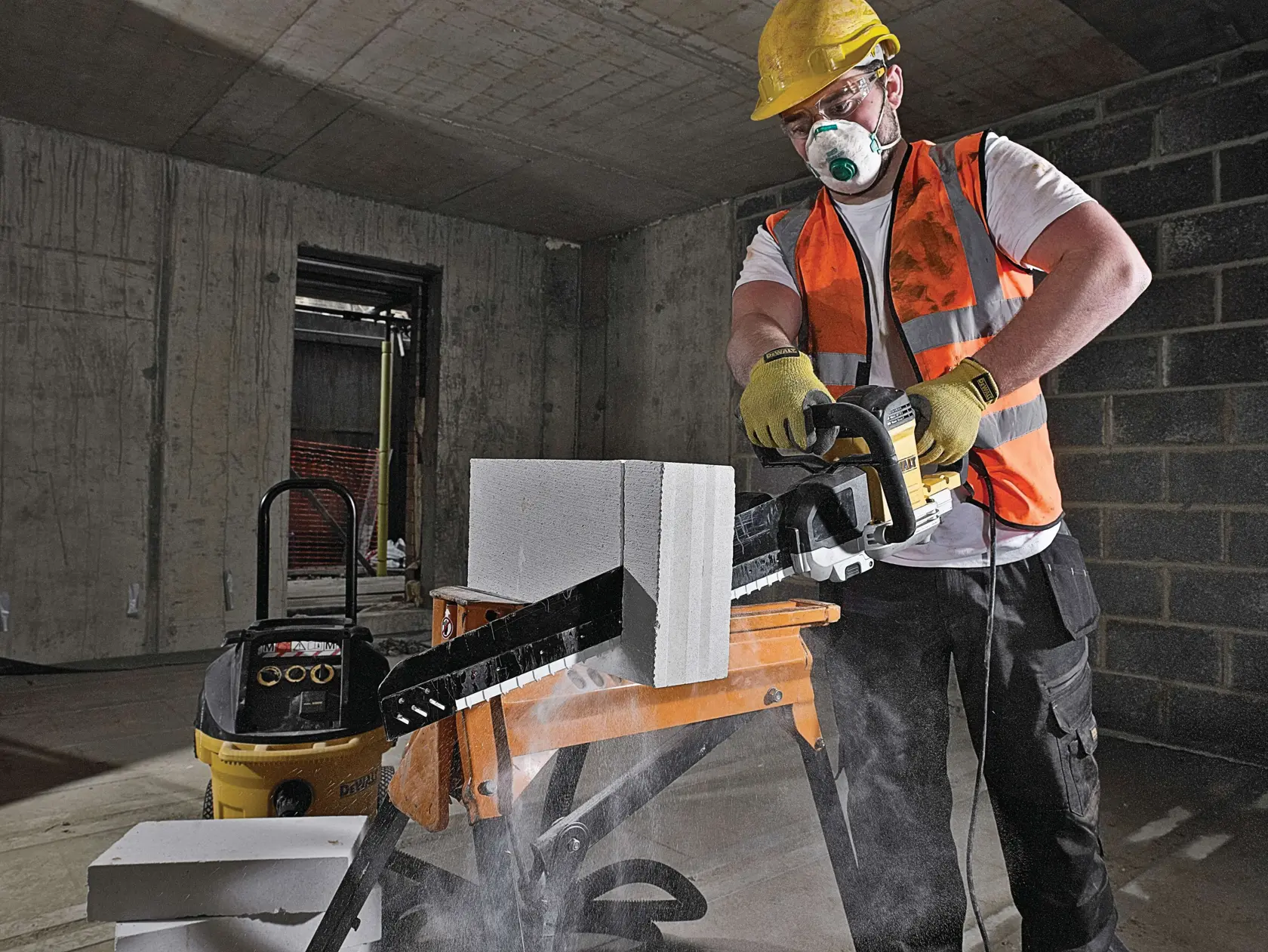Trabajador corta bloque de concreto con herramienta DEWALT en interior, lleva chaleco, guantes y casco.
