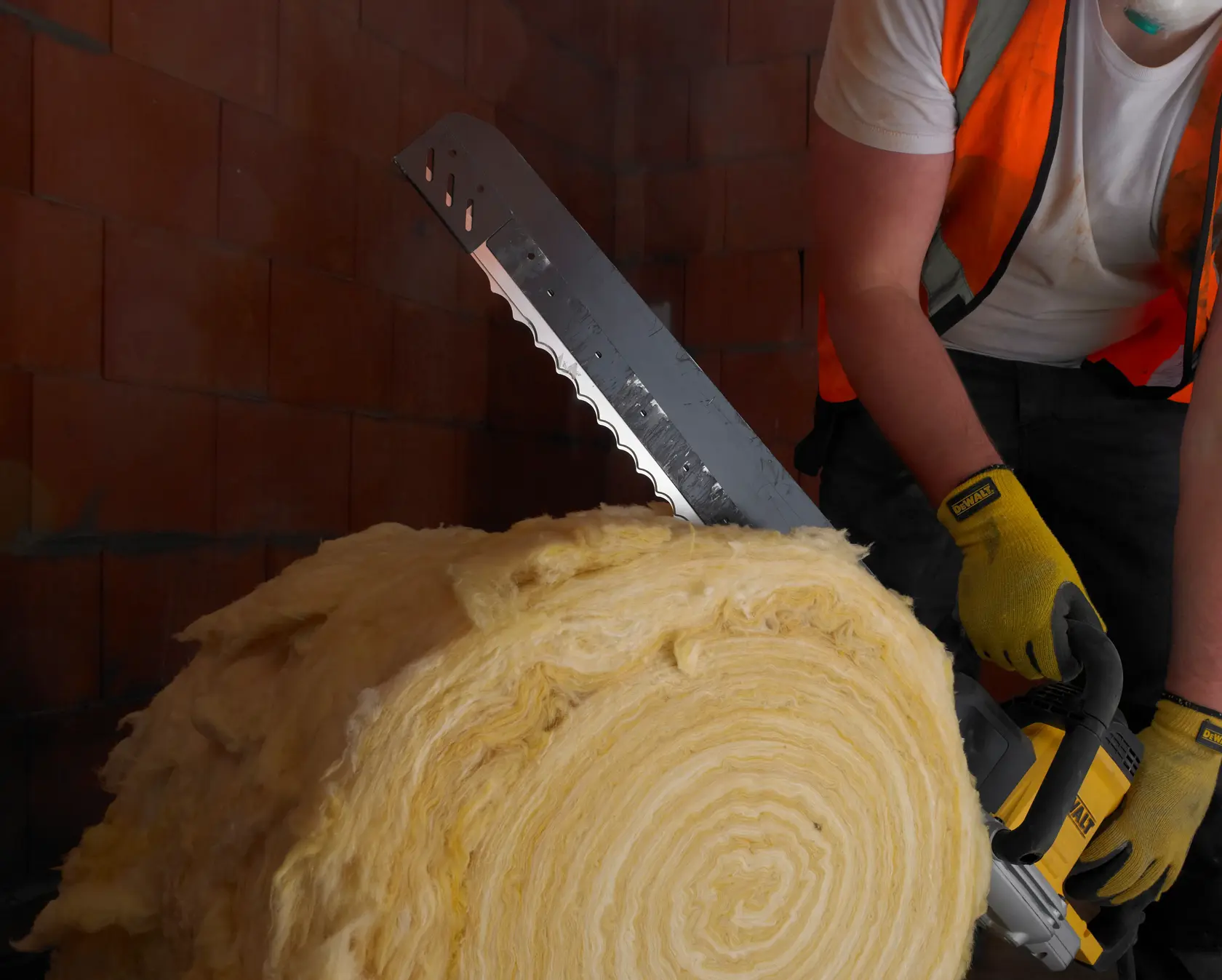 Persona corta rollo de aislamiento con sierra DEWALT, usando guantes y chaleco en zona de ladrillos.