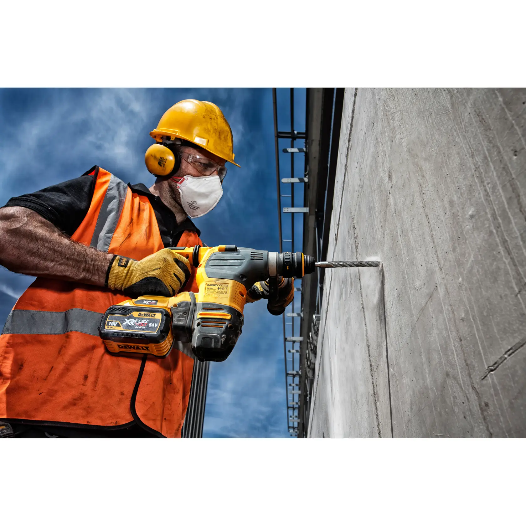 Worker in safety gear drills concrete wall with a DEWALT cordless rotary hammer under blue sky.