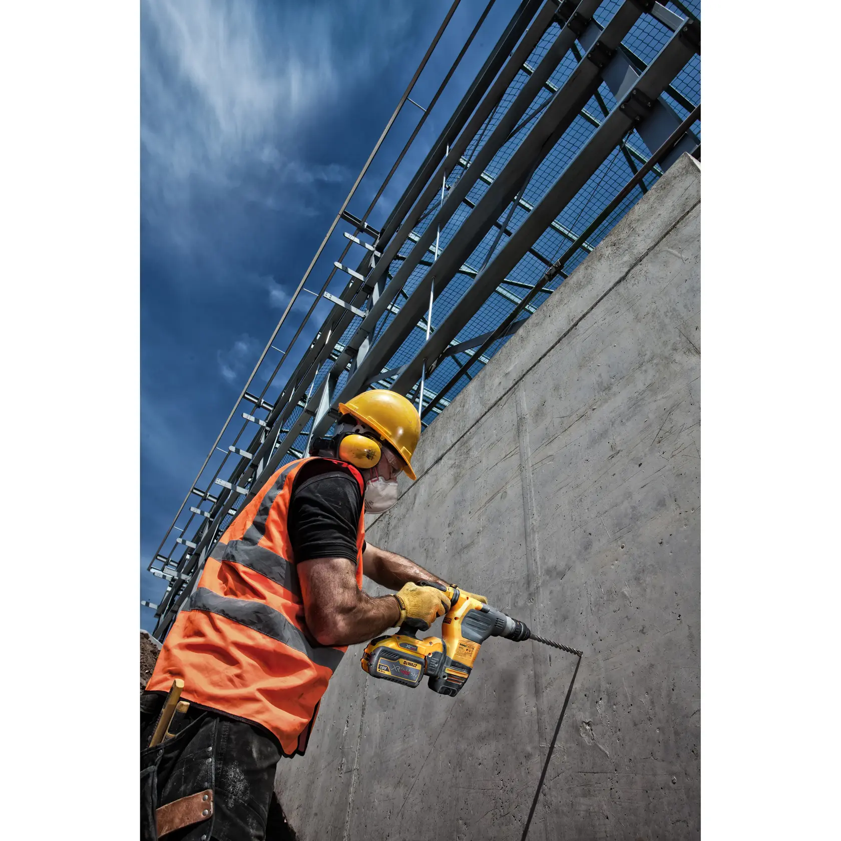 Worker uses a DEWALT drill on a concrete wall, wearing safety gear and orange vest on a construction site.