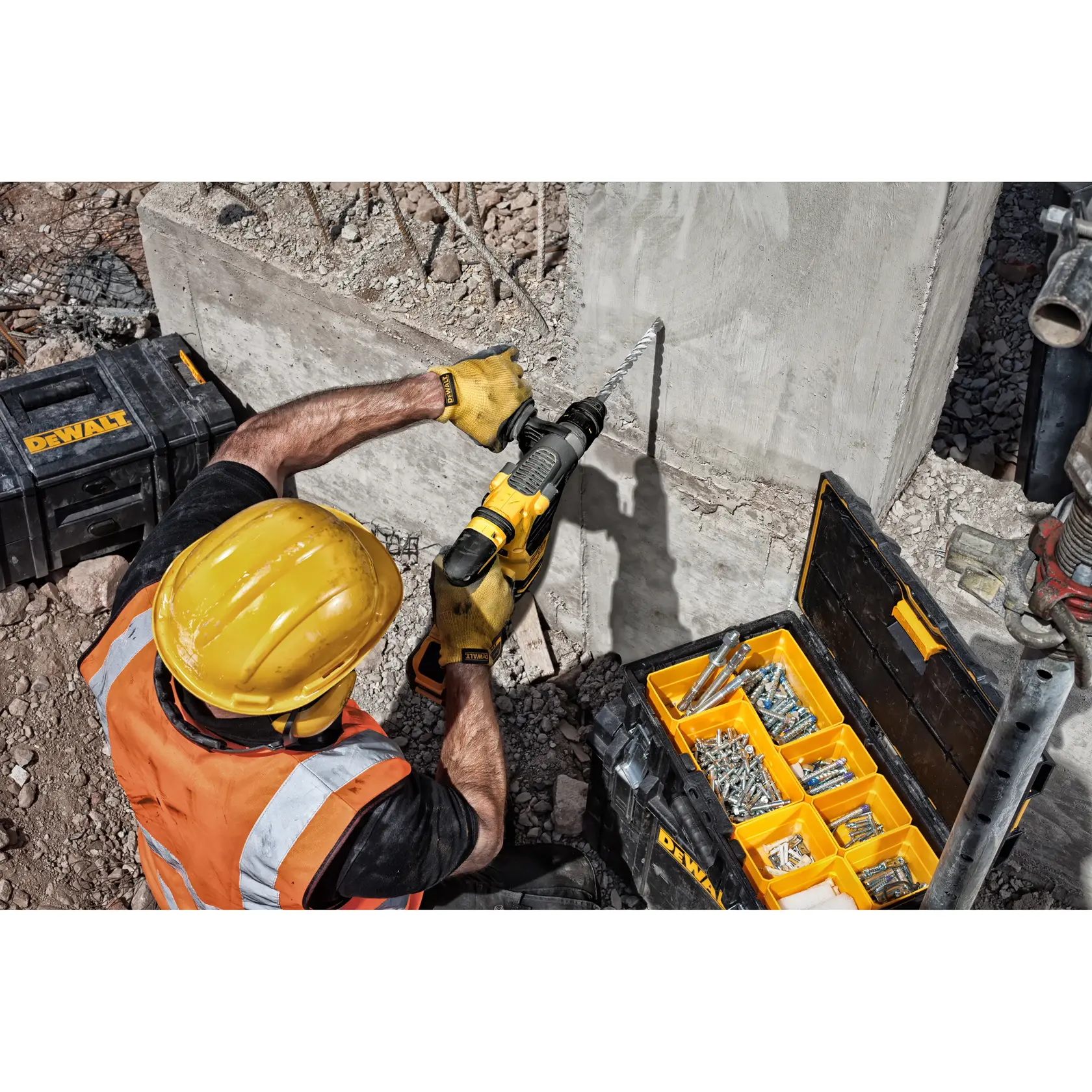 Worker drills concrete wall using DEWALT tool. Toolboxes with screws and bolts are open nearby at construction site.