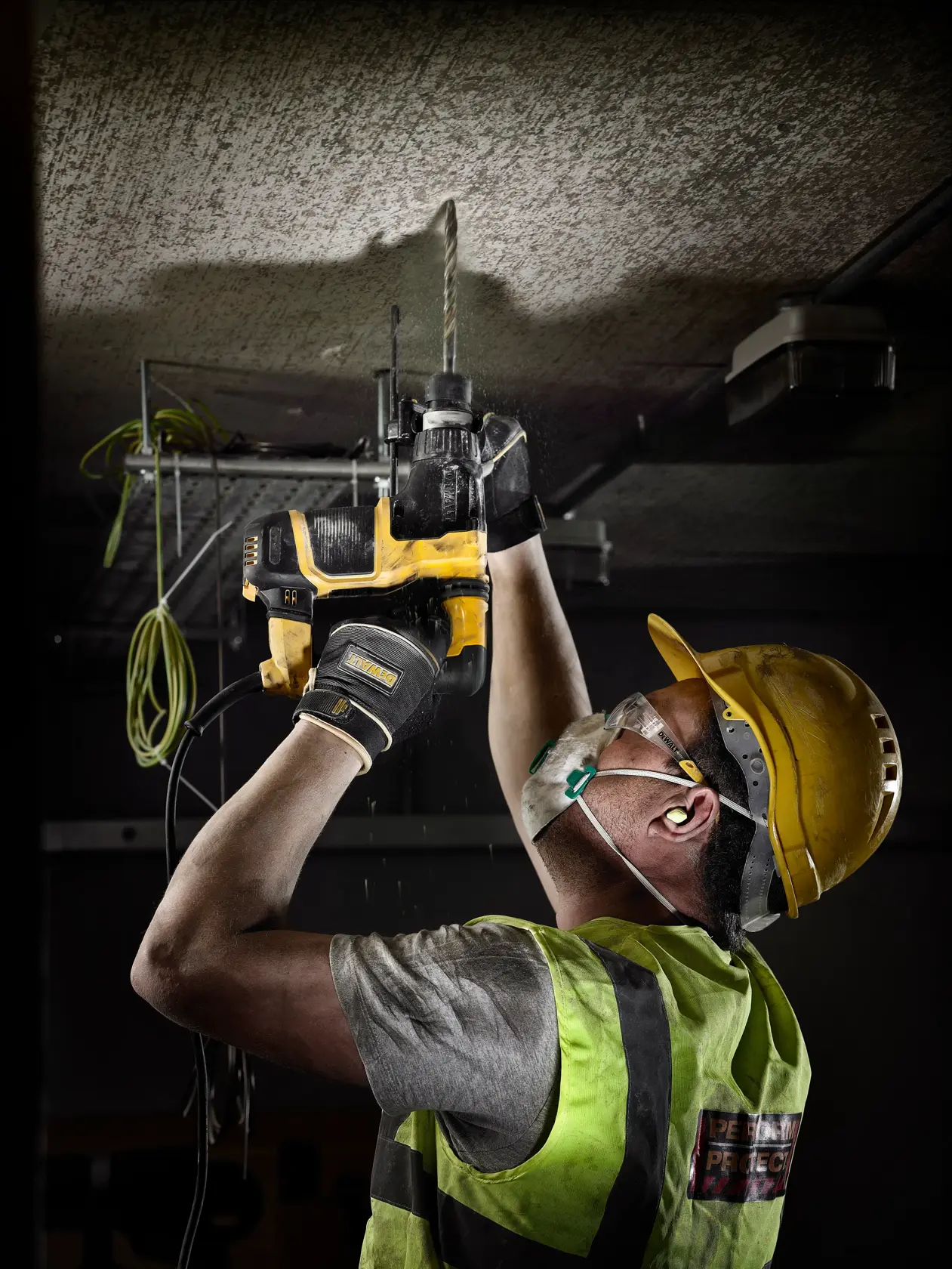 Worker using a DEWALT drill on a ceiling, wearing safety gear, gloves, helmet, and mask.