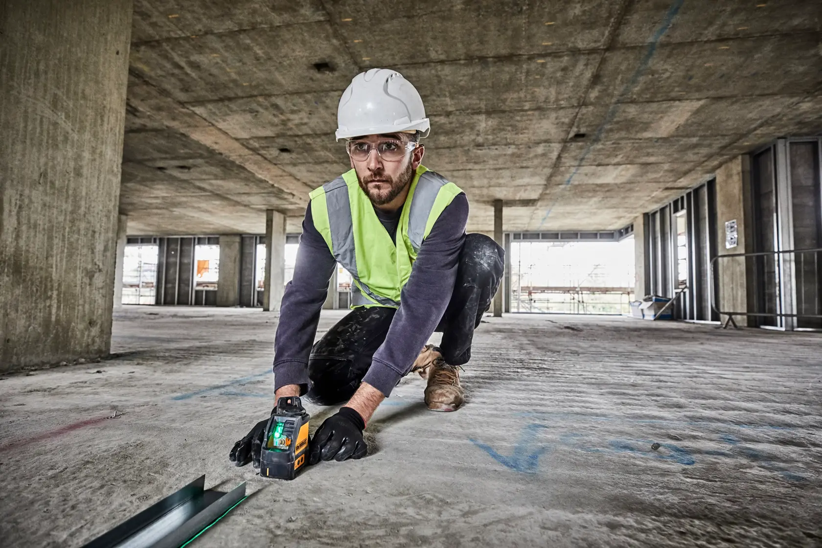 A construction worker wearing a hard hat, high-visibility vest, gloves, and boots kneels on the concrete floor of a large building under construction. The worker is using a laser measuring tool to check alignment or measurements.