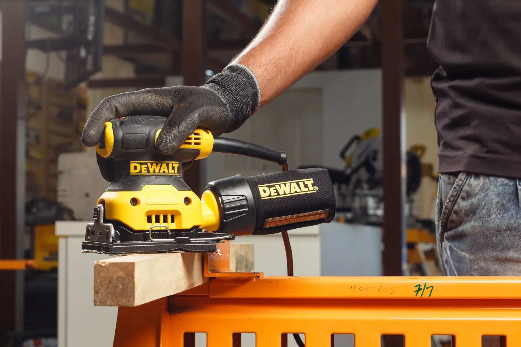 A close-up of a person using a DEWALT DWE6411 yellow and black electric sander to smooth a wooden surface outdoors. The person is wearing protective gloves.