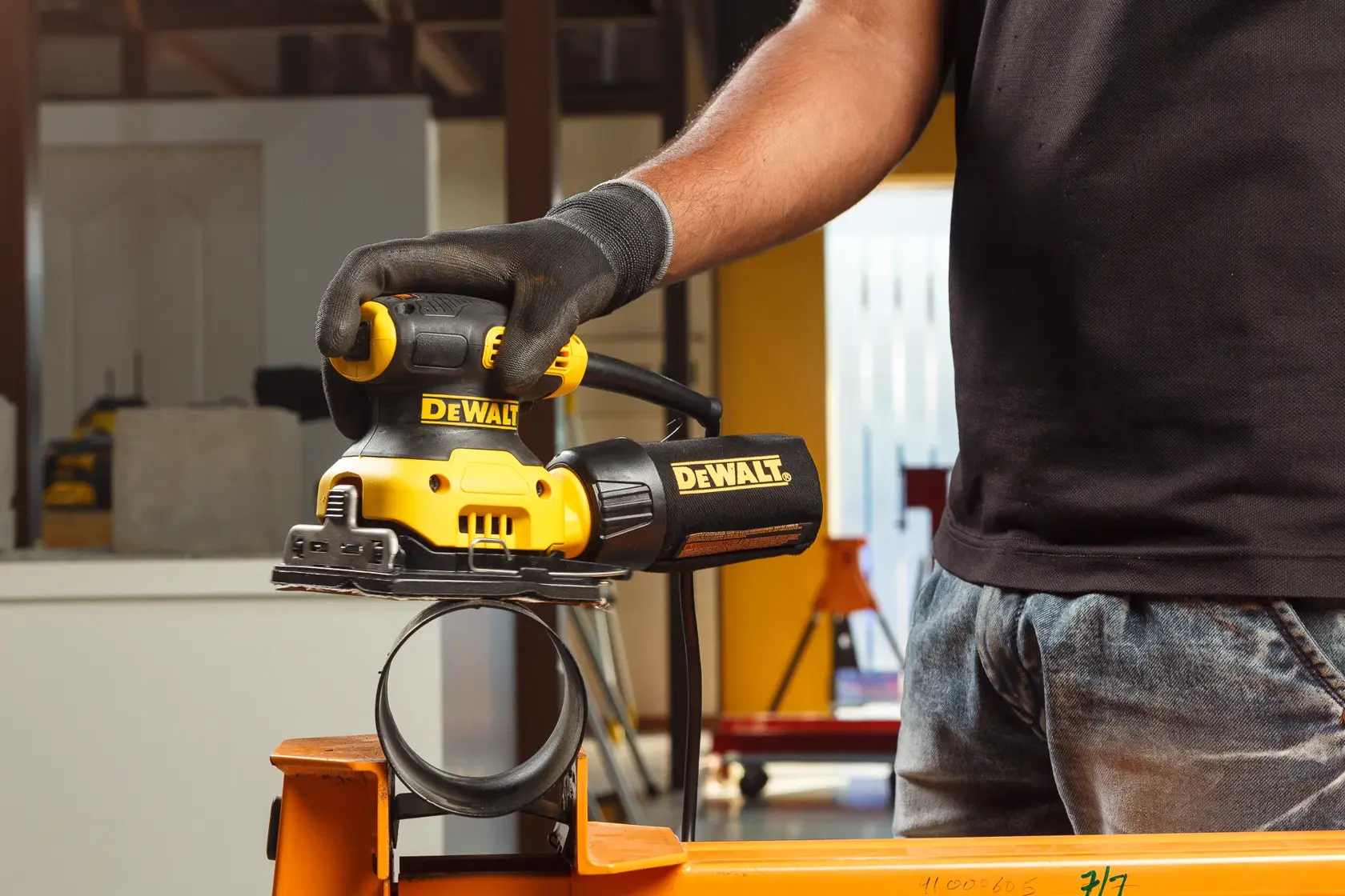 A person using a DEWALT electric sander on a wooden surface inside a workshop or garage. The sander is yellow and black, and the person's hand is holding it against the wood.
