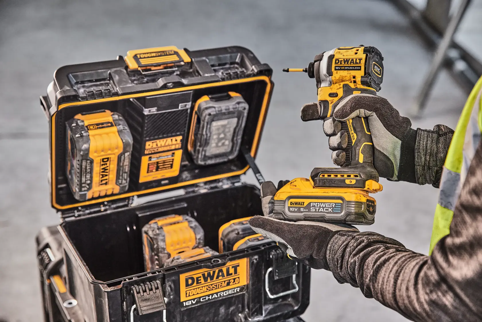 Person hält DEWALT-Bohrer, Ladekoffer mit Batterien und Werkzeugen sichtbar. Industrielles Umfeld.